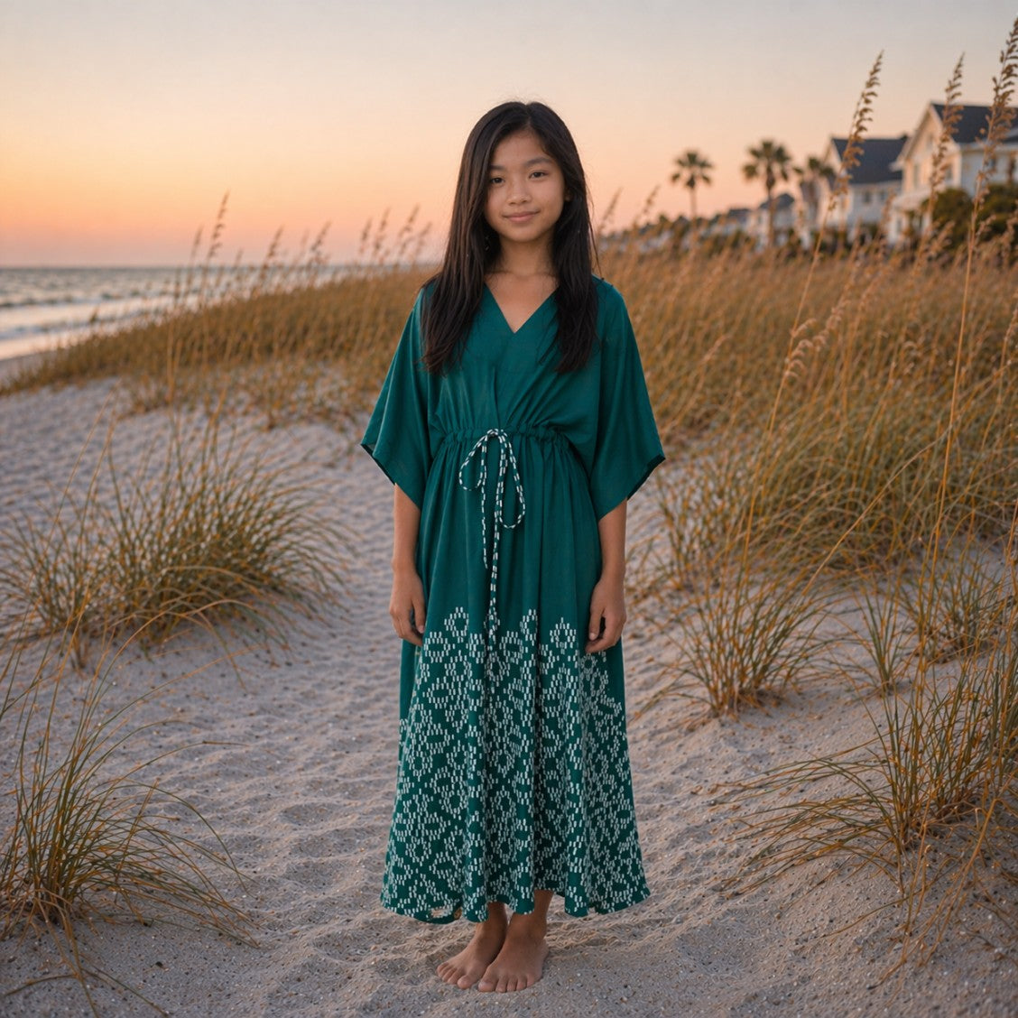 Young girl in a green dress standing on a sandy beach with grasses and houses in the background.

