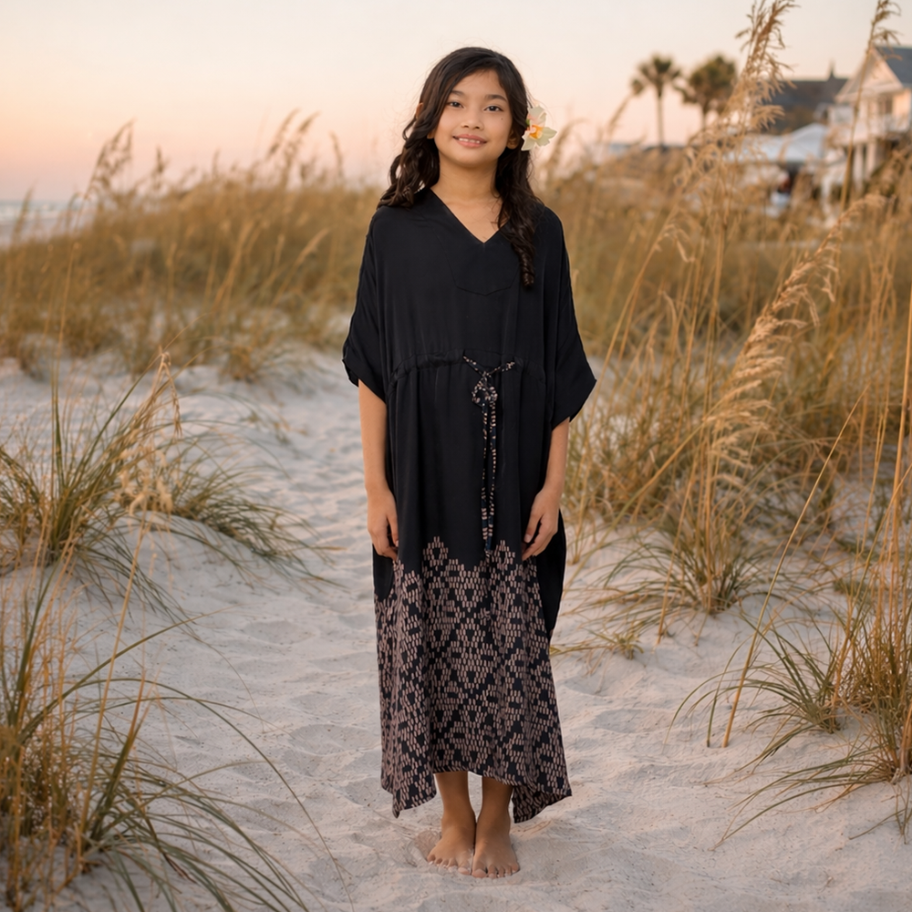 Woman in a black dress standing on a sandy beach with grasses and houses in the background.