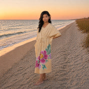 A young girl stands barefoot on a sandy beach at sunset wearing a light beige kaftan dress with a large pink and green floral print near the hem.