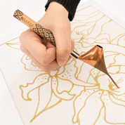 A close-up of a hand holding a Nafis brand copper calligraphy tool over white paper, drawing a gold floral pattern.