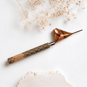 A hand tool with a copper-colored metal tip and a patterned wooden handle, photographed on a white surface with dried flowers and a stone prop.