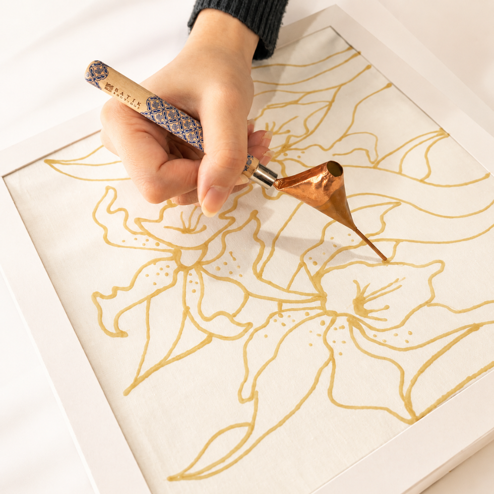 A hand holds a copper-colored metal calligraphy brush over a white sheet of paper with gold floral line art, inside a white frame.