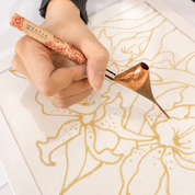 A close-up of a hand holding a Bastik Gold pen tool and applying metallic gold floral ink to a white sheet of paper.