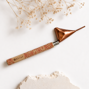 A decorative metal and wood tool with a copper-toned conical head and a patterned beige handle, styled on a white surface with dried flowers and a stone fragment.