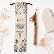 A flat-lay product photograph of a Batik Boutique bamboo-handled scraper tool next to its printed canvas drawstring pouch, styled with dried flowers and a stone on a white background.