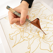 A close-up of a hand holding a RAINITE calligraphy brush over a white canvas with gold floral line art. The brush has a patterned green and beige handle and a copper-colored ferrule.