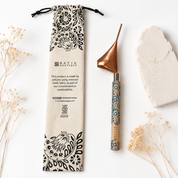 A handcrafted wooden belly scraper tool with a copper-colored metal head and a blue patterned handle, displayed next to its beige drawstring pouch with black floral prints. The set is styled on a white background with dried flowers and a stone slice.