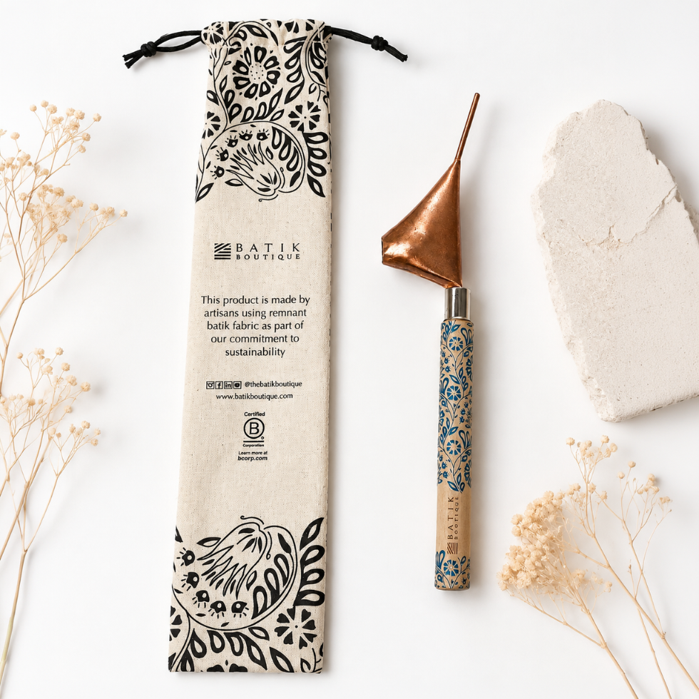 A handcrafted wooden belly scraper tool with a copper-colored metal head and a blue patterned handle, displayed next to its beige drawstring pouch with black floral prints. The set is styled on a white background with dried flowers and a stone slice.