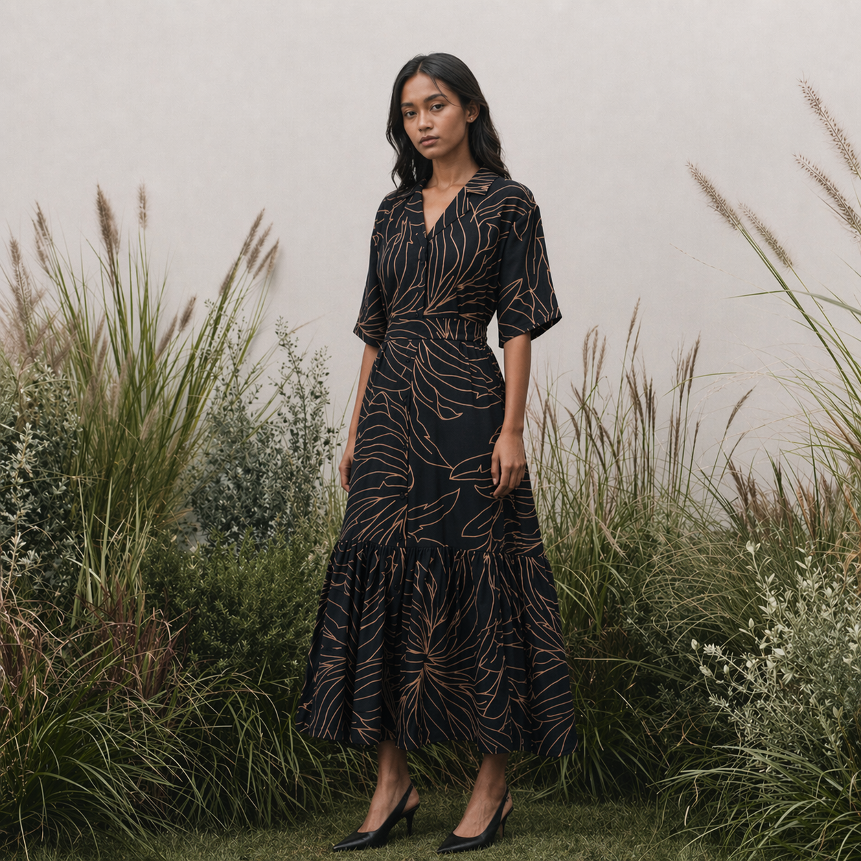 Woman in a black batik dress with gold leaf pattern standing in a natural setting with tall grasses.