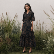 Woman in a black batik dress with gold leaf pattern standing in a natural setting with tall grasses.