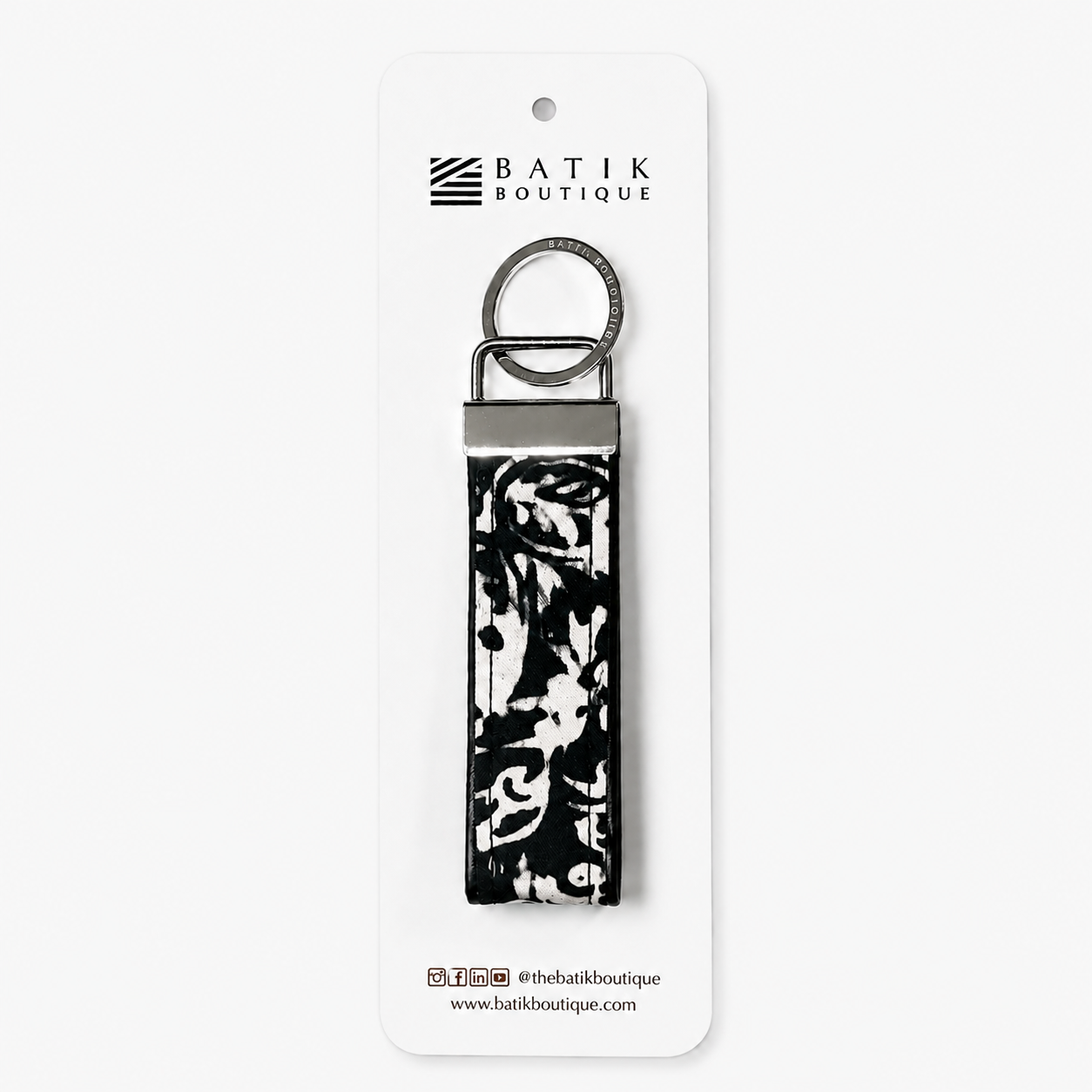 A Batik Boutique keychain with a black and white floral ikat pattern on a fabric strap, attached to a silver metal ring and hanger, displayed on a white backing card.