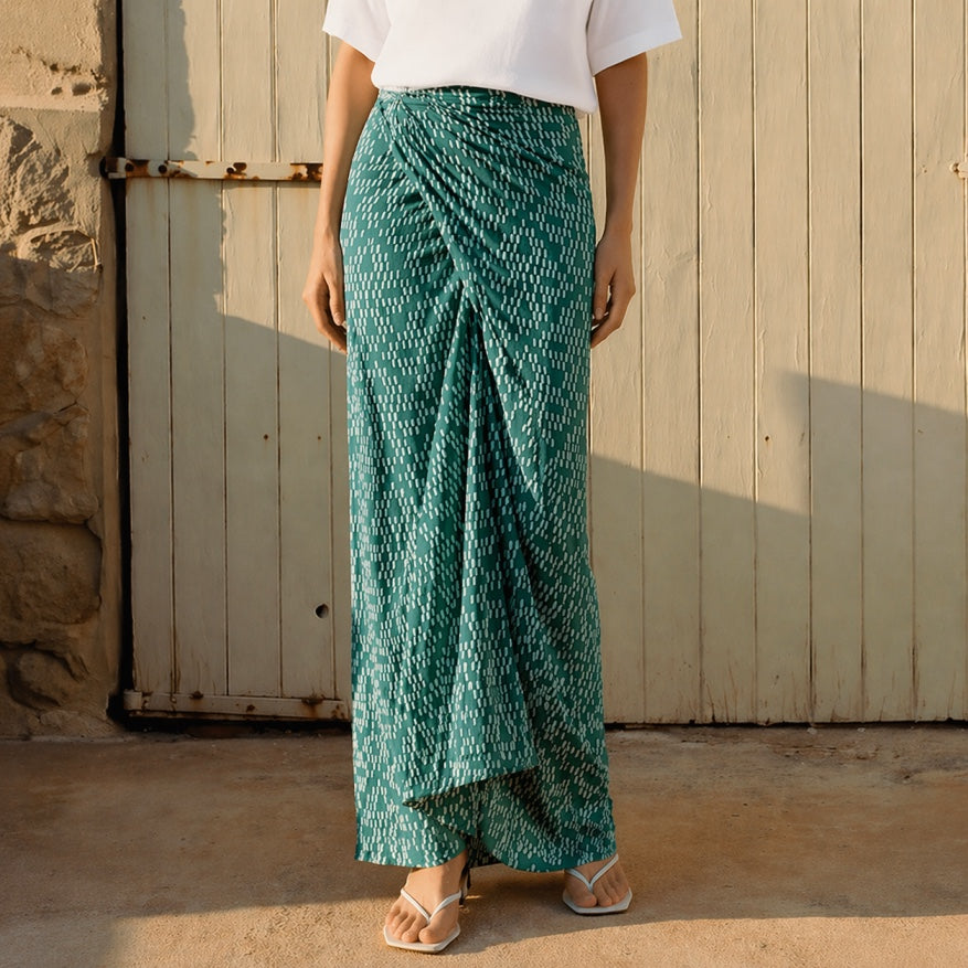 Person wearing a green patterned skirt standing against a wooden wall.