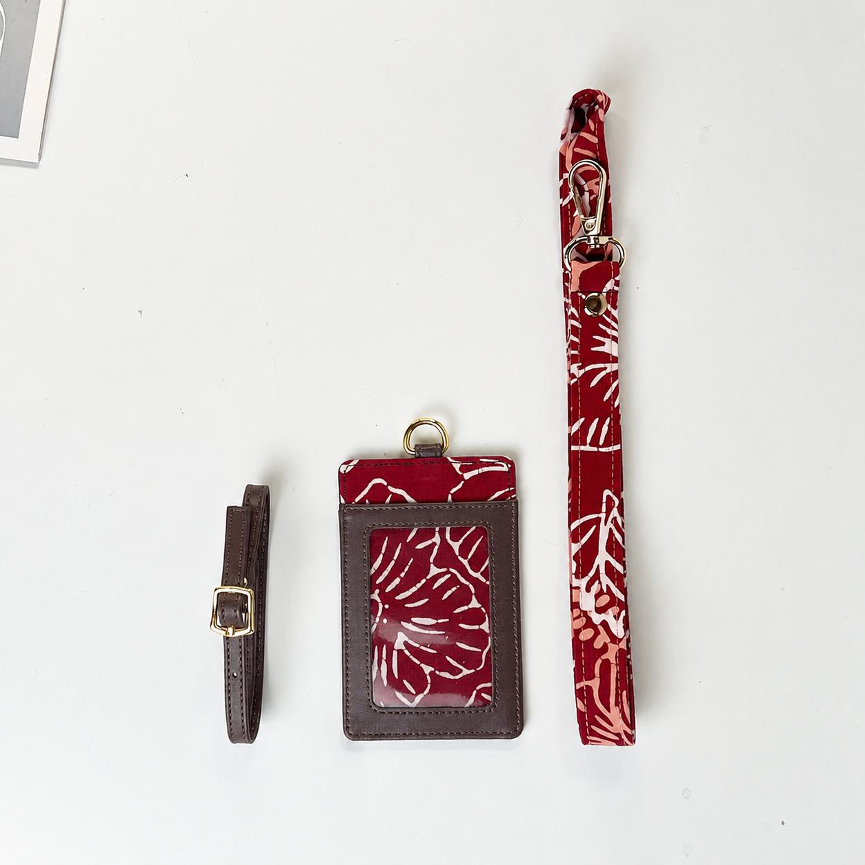 A red and white patterned card holder set with a matching wrist strap and a brown leather strap with a gold buckle, laid out on a white background.