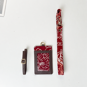 A red and white patterned card holder set with a matching wrist strap and a brown leather strap with a gold buckle, laid out on a white background.