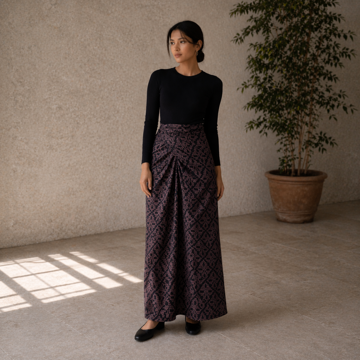 Woman wearing a black top and patterned skirt standing in a room with a plant and sunlight.
