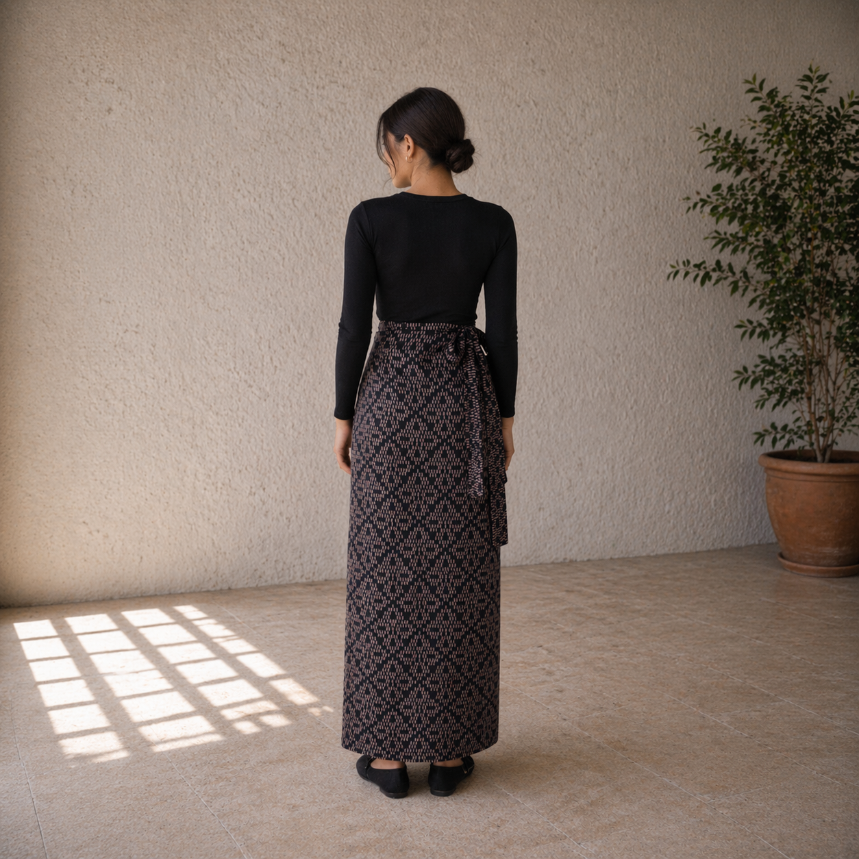 Woman wearing a black top and patterned skirt standing in a room with a plant and sunlight.