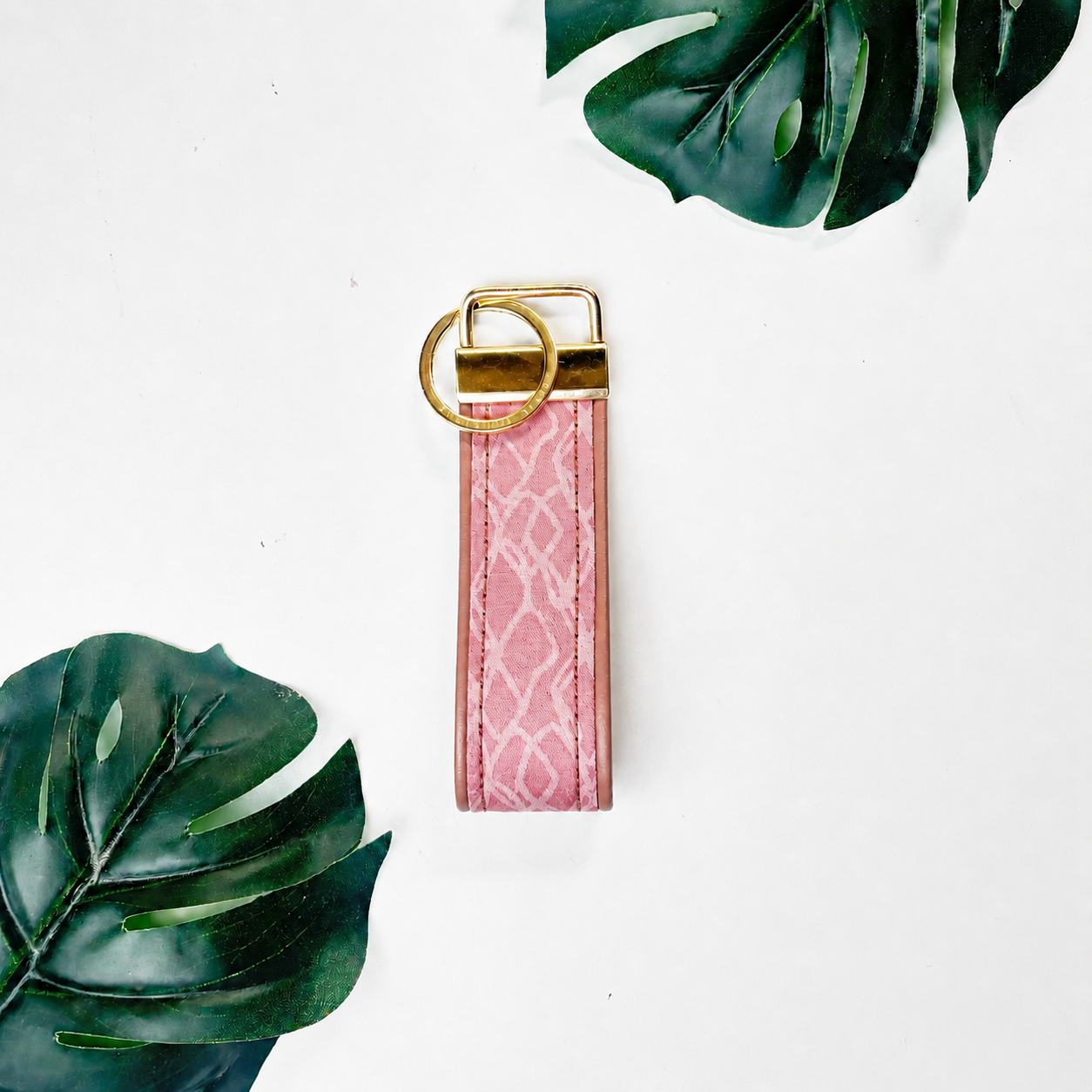 A pink faux-snakeskin leather keychain strap with a gold-tone metal key ring and split ring, displayed on a white background with decorative green tropical leaves.