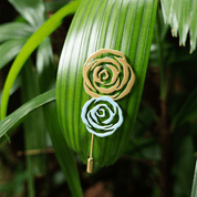 Gold and silver rose brooches pinned to a green tropical palm leaf, showcasing their contrast and intricate rose petal design.