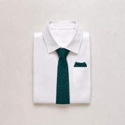 A folded white dress shirt with a dark green patterned necktie and matching pocket square, arranged on a light beige background.
