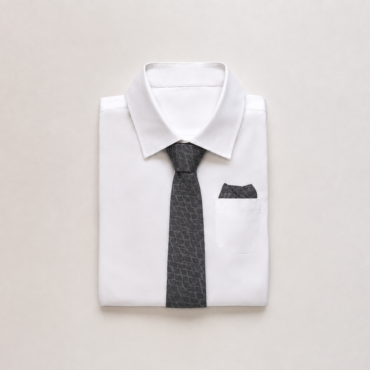 A folded white dress shirt with a dark gray patterned necktie and matching pocket square, arranged on a light beige background.