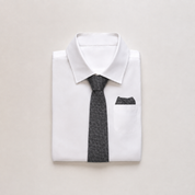 A folded white dress shirt with a dark gray patterned necktie and matching pocket square, arranged on a light beige background.