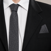A close-up of a men's formal outfit featuring a dark gray patterned silk tie and matching pocket square, styled with a white dress shirt and a black suit jacket.