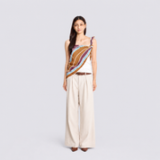 A woman stands against a light gray studio background wearing a sleeveless wrap-style top with diagonal striped panels in white, orange, magenta, and burgundy, paired with wide-leg beige trousers and brown pointed shoes.