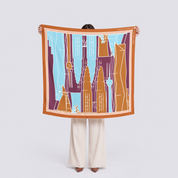 A person stands against a light gray studio background holding up a square scarf. The scarf features a stylized cityscape illustration with orange, magenta, light blue, and white elements, framed by a bright orange border.