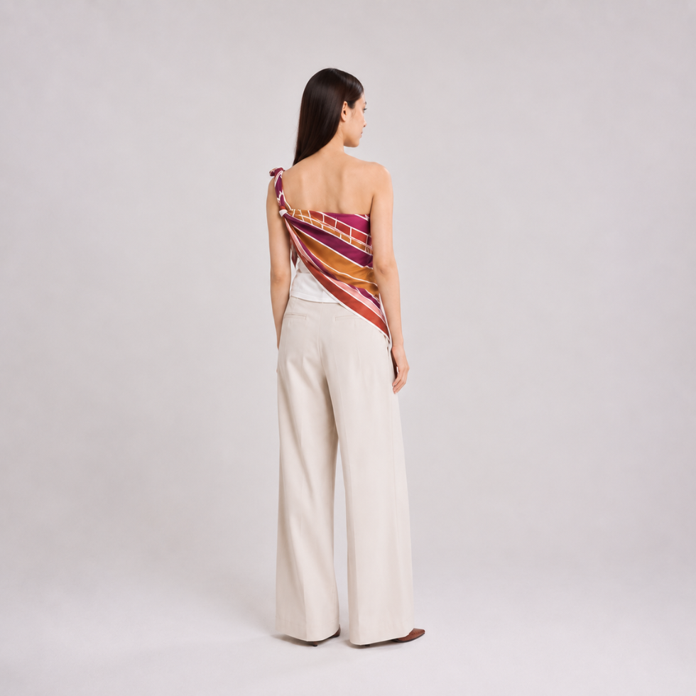A woman stands with her back to the camera wearing a one-shoulder wrap top with a colorful striped pattern in red, orange, yellow, and purple, paired with high-waisted cream wide-leg trousers.