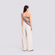 A woman stands with her back to the camera wearing a one-shoulder wrap top with a colorful striped pattern in red, orange, yellow, and purple, paired with high-waisted cream wide-leg trousers.