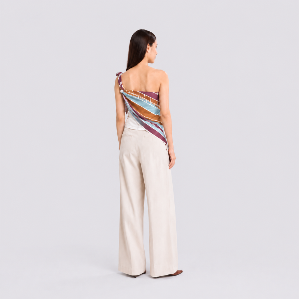 A woman stands with her back to the camera wearing a one-shoulder wrap top with a colorful striped pattern in red, orange, yellow, and purple, paired with high-waisted cream wide-leg trousers.