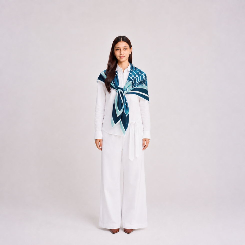 A woman stands against a light gray studio background wearing a white long-sleeve top and wide-leg white pants, with a patterned scarf draped around her shoulders in shades of blue, teal, and navy.