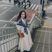 Woman in a white dress with a blue scarf standing on a city street.