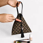 A black and gold patterned handbag with a tassel zipper is held by two hands, with cosmetic items placed near the bottom of the frame on a light gray background.