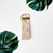 A vertical leather keychain strap in a dusty pink color with a white leaf pattern, featuring a gold metal ring and loop, displayed on a white background with green monstera leaves.