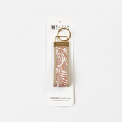 A Batik Boutique keychain featuring a rectangular pink and white patterned fabric strap with a brass metal ring and connector, mounted on a white cardboard display card.