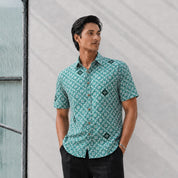 Man wearing a patterned teal shirt standing against a light gray wall.