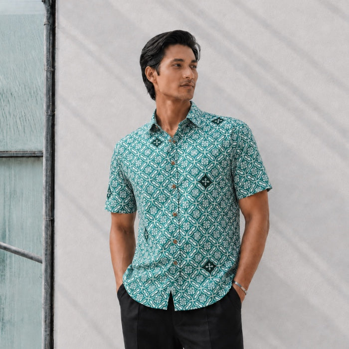 Man wearing a patterned teal shirt standing against a light gray wall.
