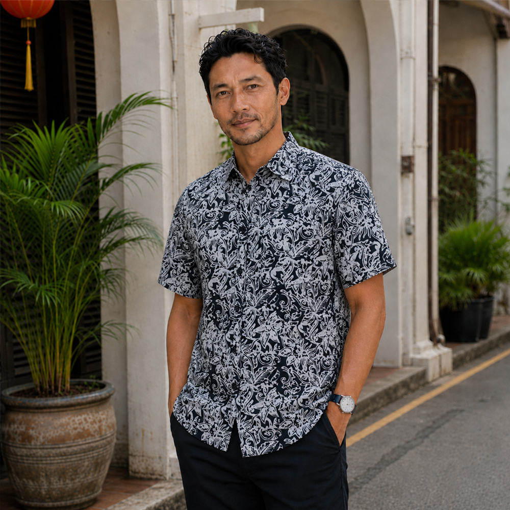 A man wearing a short-sleeve button-up shirt with a black and white botanical pattern, standing outdoors in front of a colonial-style building with potted plants.
