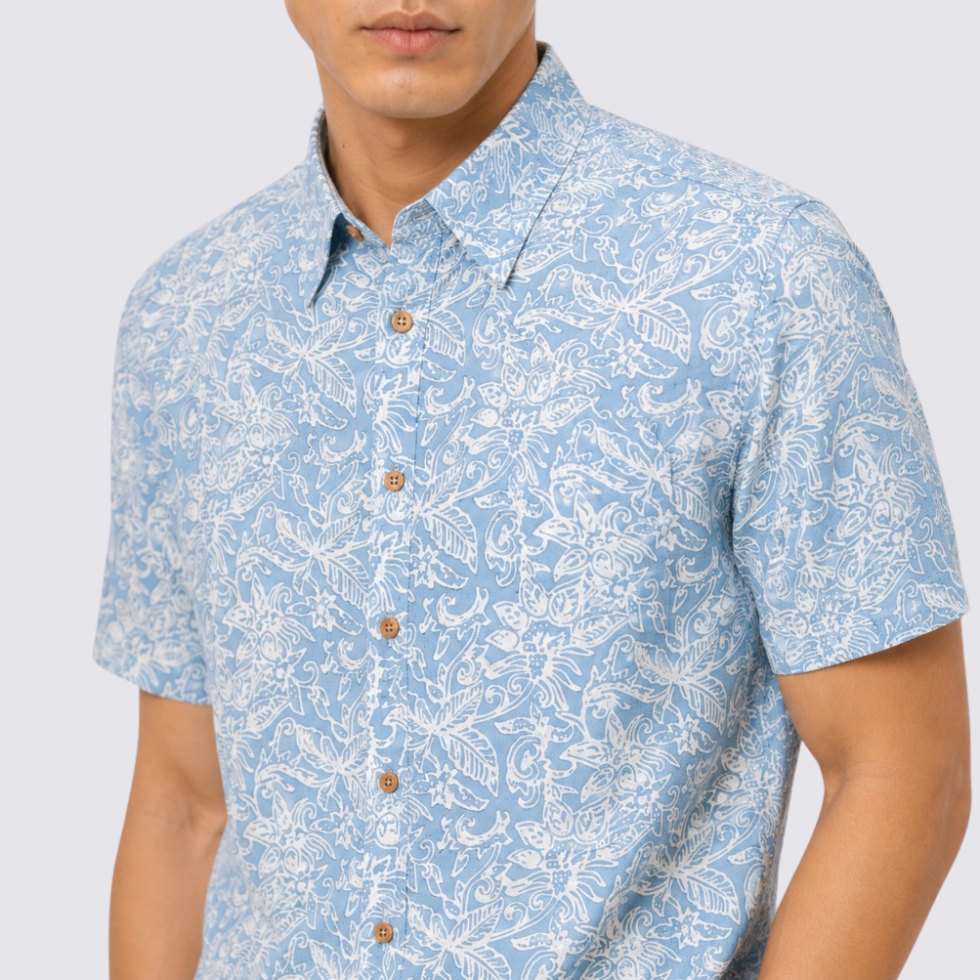 A man wearing a light blue short-sleeve button-up shirt with a white tropical floral and leaf pattern, featuring brown buttons.