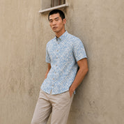 A man wearing a blue patterned batik shirt and beige pants is leaning against a beige wall.