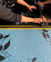 Close-up of a fabric with leaf pattern being measured by hands using a yellow measuring tape.