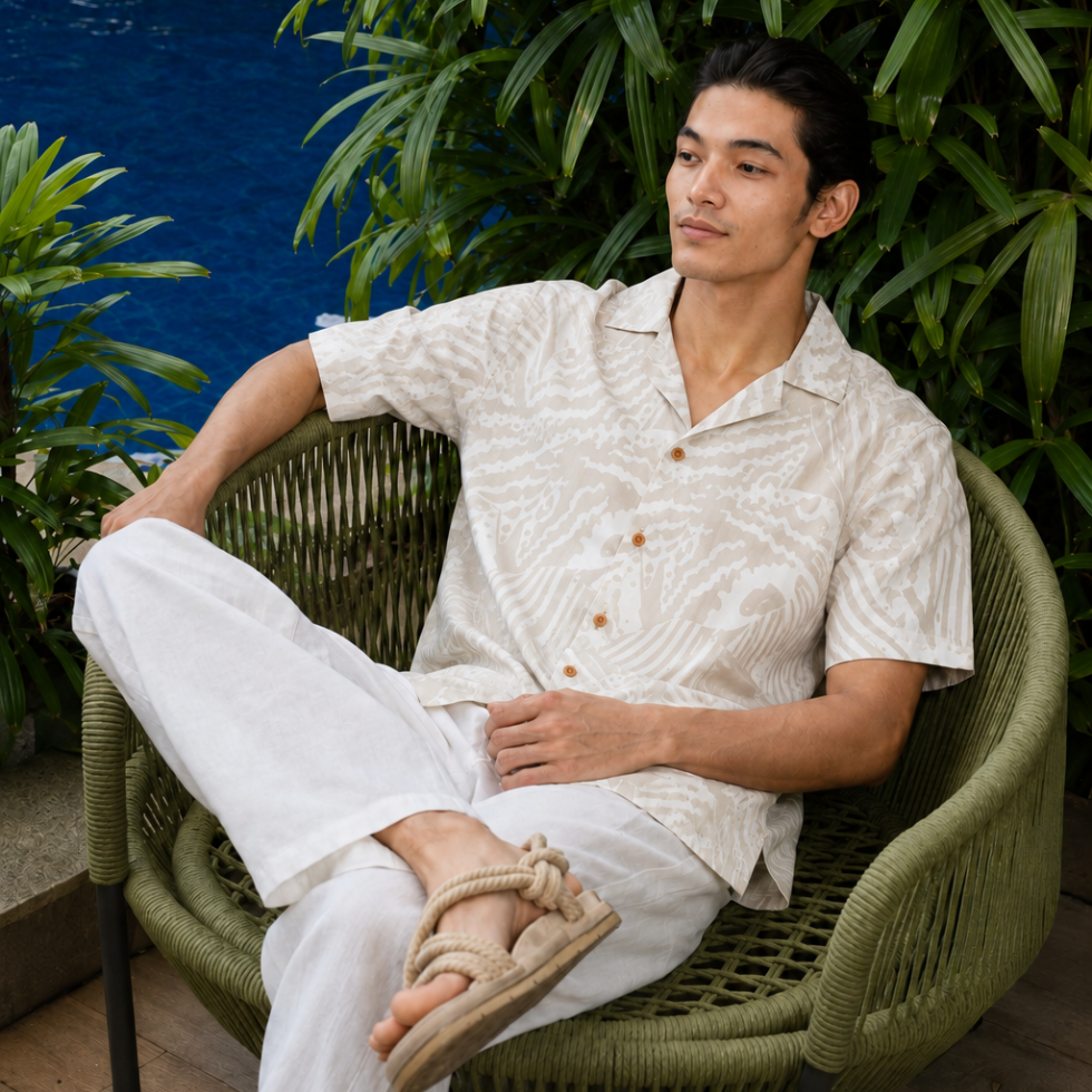 Man in batik shirt with gray printed patterns sitting on a chair with greenery behind and by the pool