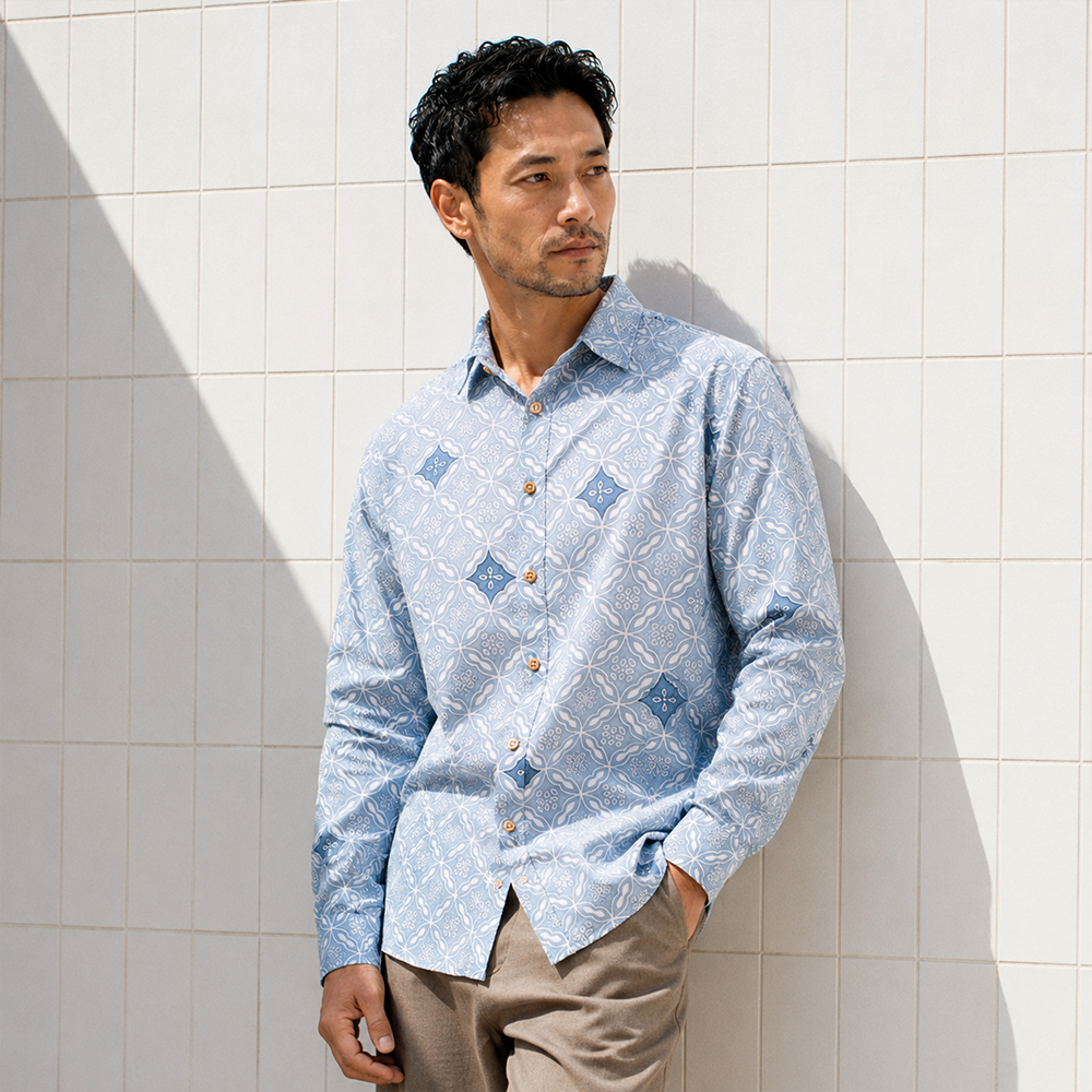A man wearing a light blue long-sleeve button-up shirt with a white geometric pattern and blue diamond motifs, paired with beige trousers, standing against a white tiled wall.