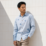 A man wearing a light blue long-sleeve button-up shirt with a white geometric pattern and blue diamond motifs, paired with beige trousers, standing against a white tiled wall.