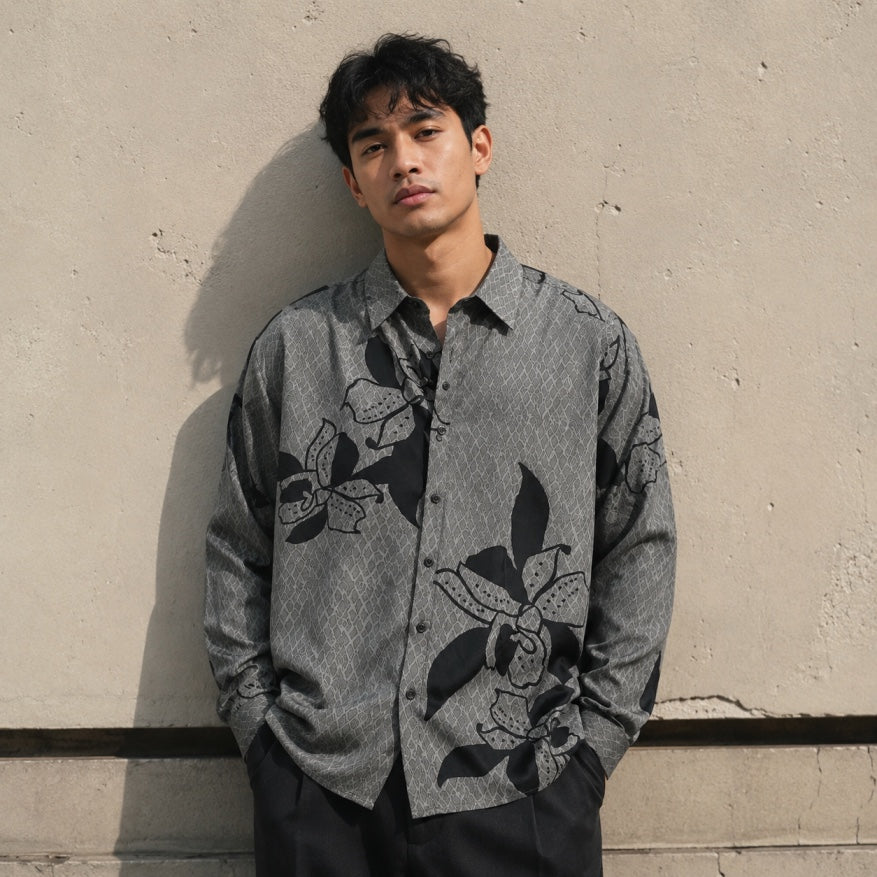 A man wearing a gray long-sleeve button-up shirt with a black floral pattern, leaning against a textured concrete wall.
