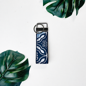 A vertical leather keychain strap in a blue batik with white patterns pattern, featuring a silver metal ring and loop, displayed on a white background with green monstera leaves.