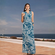 A woman stands outdoors by the ocean wearing a sleeveless maxi dress with a bold blue and white abstract pattern. The dress has a gathered waist and side pockets.