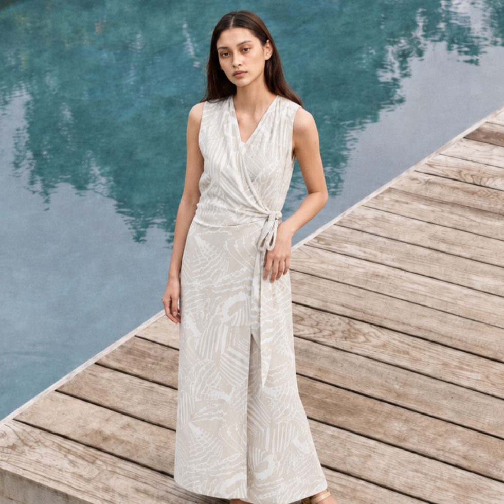 A woman stands on a wooden dock beside a swimming pool wearing a sleeveless, wrap-style maxi dress with a side tie and a light beige leaf pattern.