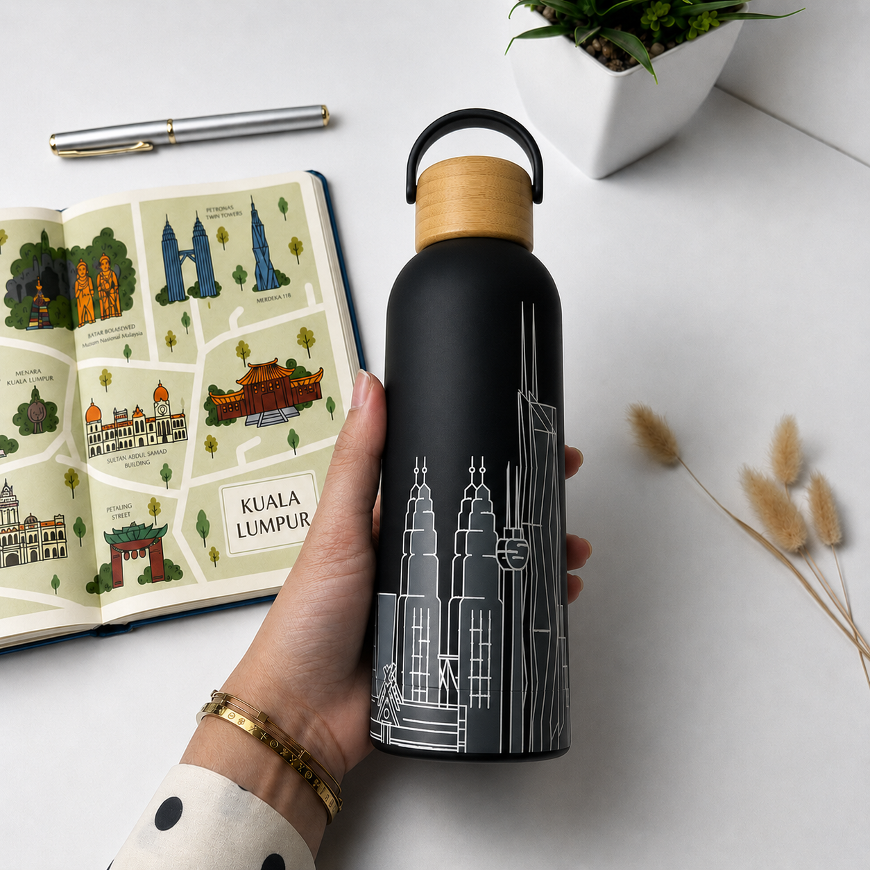 A hand holds a matte black water bottle with a light wood cap and white Kuala Lumpur cityscape line art. An illustrated open book, a pen, and dried stems are arranged on a white surface.